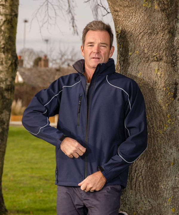 A man wearing a navy blue Result R118A jacket stands outdoors, leaning against a tree. He is zipping up his jacket, with trees and a grassy area in the background.