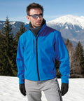 A person wearing a blue jacket, gray scarf, gray pants, black gloves, and Result R121A sunglasses stands in a snowy mountainous area. Pine trees and snow-capped peaks are visible in the background.