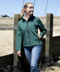A person wearing a green Result R121F jacket and blue jeans is standing on a pebbled beach leaning against a wooden structure, with boats in the background.