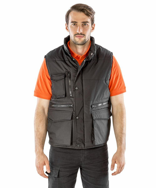 A man wearing a black Result R127A utility vest over an orange polo shirt stands facing forward with a neutral expression.