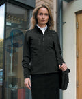 A woman stands outside a building, wearing a black jacket and a black skirt, holding a black Result R128F shoulder bag.