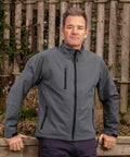 A man in a Result R128X jacket stands outdoors in front of a wooden fence, looking directly at the camera with his hands resting on a ledge.