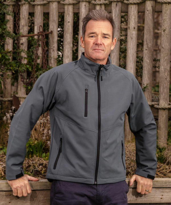 A man in a Result R128X jacket stands outdoors in front of a wooden fence, looking directly at the camera with his hands resting on a ledge.