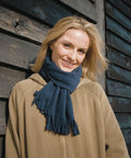 Smiling person with blonde hair wearing a Result R143X tan jacket and blue scarf, standing against a weathered wooden wall.