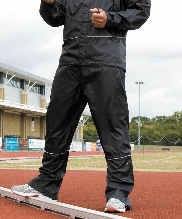 A person standing on a track wearing a black R156A tracksuit and sports shoes from Result. The background shows a building and some trees.