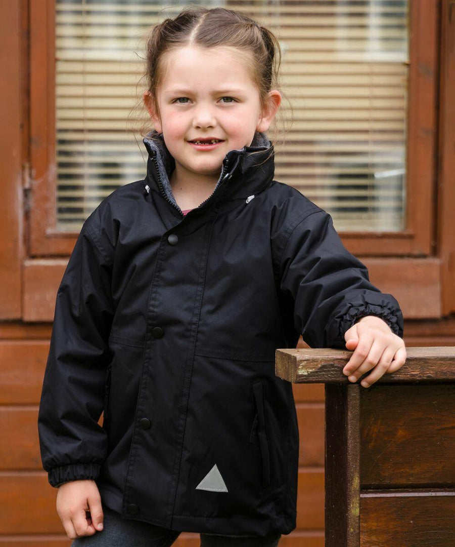 A young girl with pigtails stands outdoors wearing a black R160J jacket from Result. She is leaning against a wooden railing with a building and window blinds in the background.