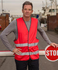 A man in a red Result R200X reflective safety vest stands by a barrier gate with a "STOP" sign on it in an outdoor setting with boats and buildings in the background.