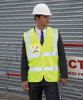 Person in a suit wearing a bright yellow Result R202X reflective vest and white hard hat stands in front of a corrugated metal door, holding a clipboard.