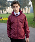 A young boy wearing a Result R203J jacket and a striped tie stands outside on a paved path, with trees and buildings in the background.