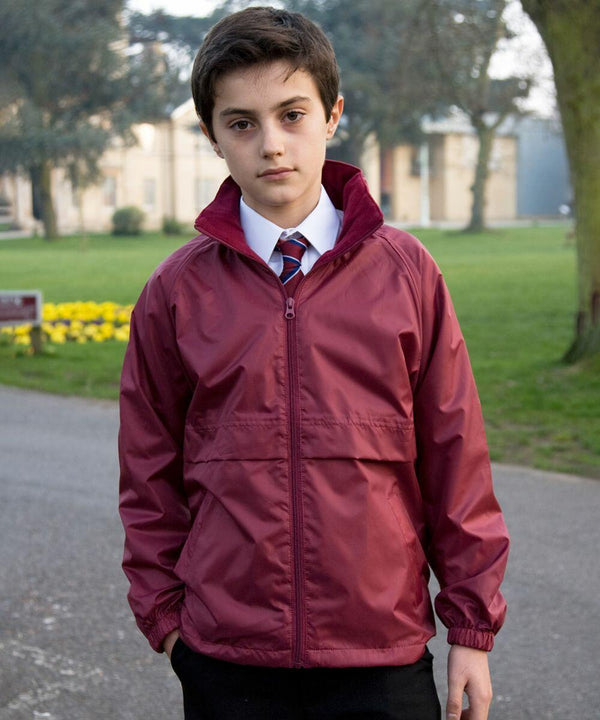 A young boy wearing a Result R203J jacket and a striped tie stands outside on a paved path, with trees and buildings in the background.