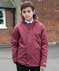 A young person stands outdoors wearing a Result R207J over a school uniform in front of a brick building.
