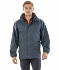 A person standing, wearing a dark blue R215X hooded jacket with front zipper and pockets from Result, looking forward.