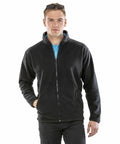 A man with sunglasses on his head is wearing a blue shirt under a zipped-up black Result R220X fleece jacket and black pants, with his left hand in a jacket pocket.