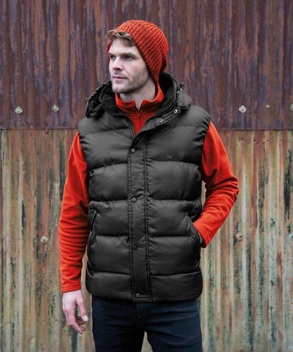 A man wearing a red knit cap, red sweater, and black puffer vest stands in front of a rusty, corrugated metal wall. He is dressed in the Result R223X.