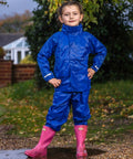 A child in a bright blue Result R225J and pink boots stands confidently outdoors on a gravel path, with a house and foliage visible in the background.