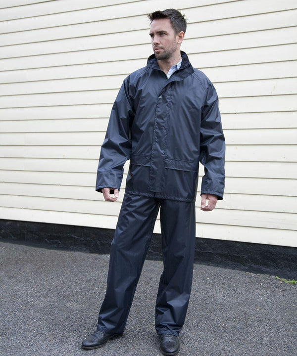 A person wearing a dark gray Result R225X rain jacket and pants stands on a wet pavement in front of a beige sided building.