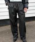Person wearing Result R226X shiny black rain pants and matching jacket, standing on a paved surface in front of a white building.