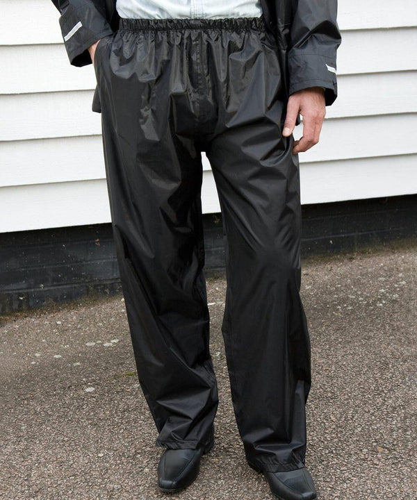 Person wearing Result R226X shiny black rain pants and matching jacket, standing on a paved surface in front of a white building.