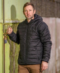 A man in a black Result R233M quilted jacket stands holding a latch on a green door, with a padlock visible near his hand. He has short hair and is looking directly at the camera.