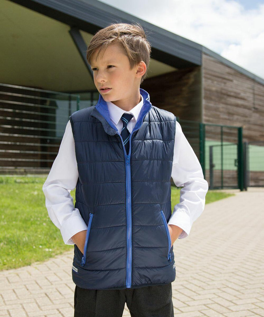 A young boy stands outdoors, wearing a white shirt, tie, and navy blue Result R234J puffer vest, with hands in his vest pockets. He is in front of a modern building on a bright, sunny day.