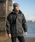 A man wearing a gray beanie, a high-collared black Result R236X jacket, and a gray scarf stands outdoors near water with a wooden building in the background.