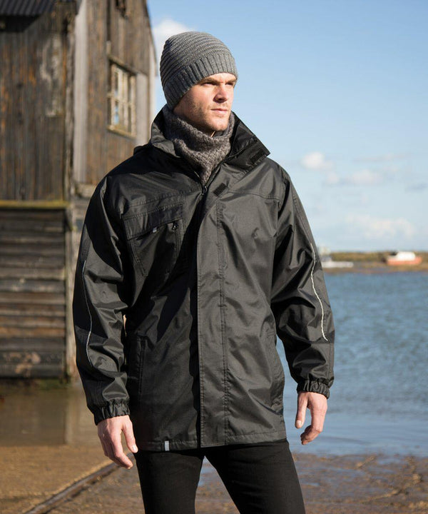 A man wearing a gray beanie, a high-collared black Result R236X jacket, and a gray scarf stands outdoors near water with a wooden building in the background.