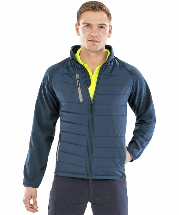 A man stands wearing a Result Genuine Recycled R237X navy blue padded jacket with a front zipper and a chest pocket. He has light brown hair and is looking directly at the camera.