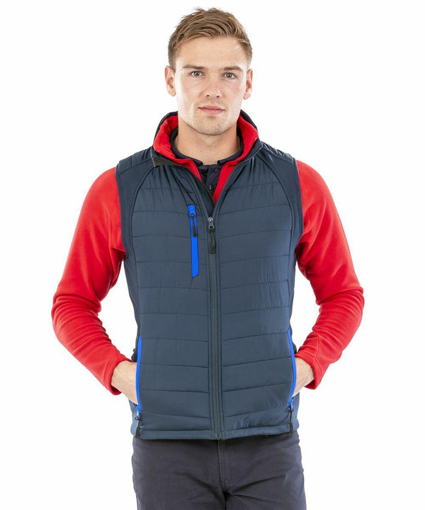 Young man wearing a red long-sleeve shirt and a dark blue Result Genuine Recycled R238X stands facing the camera with his hands in the pockets of his vest.
