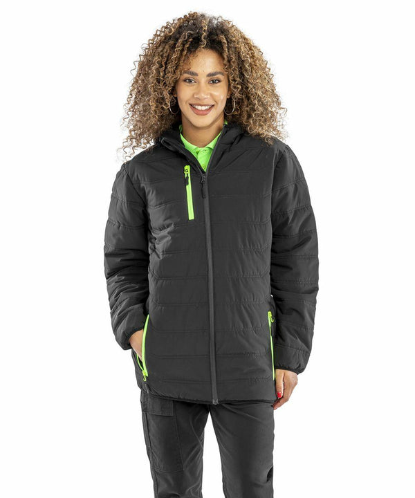 A person with curly hair wearing a black puffer jacket with neon green zipper details, the R240X by Result, stands smiling with hands in pockets.
