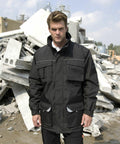A man wearing a black Result R301X utility jacket stands in front of a pile of concrete debris outdoors.