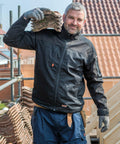 A man in work attire and gloves is carrying a stack of Result R302X roof tiles on his shoulder at a construction site. He is smiling and standing next to scaffolding and wooden framing.