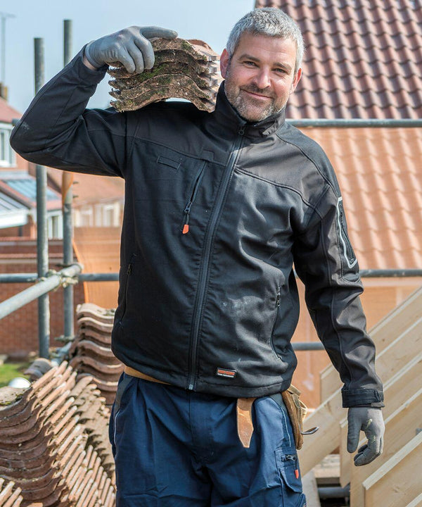 A man in work attire and gloves is carrying a stack of Result R302X roof tiles on his shoulder at a construction site. He is smiling and standing next to scaffolding and wooden framing.