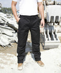 A person wearing black cargo pants and brown Result R303X work boots stands near heavy construction machinery on a muddy, debris-filled construction site.