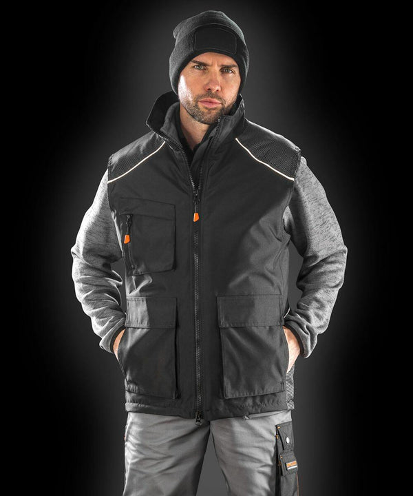 A man wearing a black beanie, a black Result R306X zip-up vest over a grey hooded sweatshirt, and grey pants stands with his hands in his pockets against a dark background.