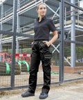 A person wearing black Result R308F stands with hands in pockets in front of a metal fence with a construction site in the background.