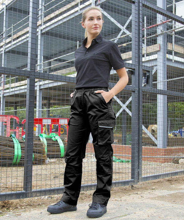 A person wearing black Result R308F stands with hands in pockets in front of a metal fence with a construction site in the background.