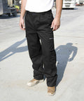 A person in Result R308X black work pants and brown work boots stands on a concrete surface with vehicles and industrial equipment in the background.
