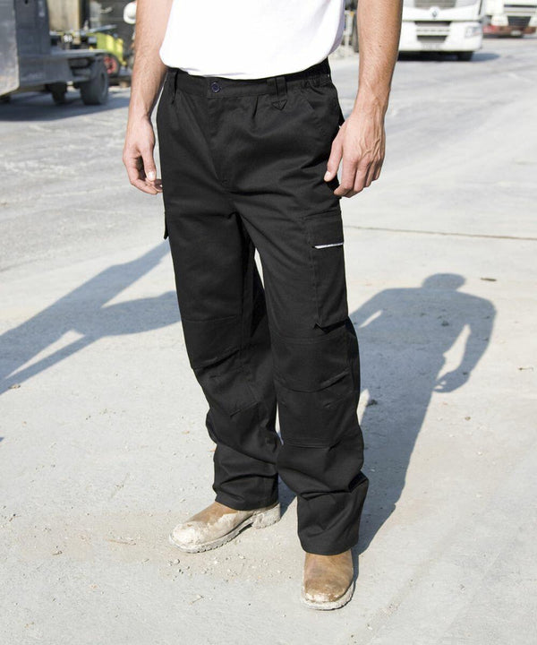 A person in Result R308X black work pants and brown work boots stands on a concrete surface with vehicles and industrial equipment in the background.