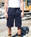 A person wearing a white shirt, navy cargo shorts, and brown Result R309X work boots stands on a construction site with heavy machinery in the background.