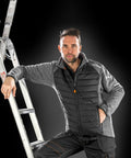 A person with short hair leans against a ladder, wearing a Result R314X gray and black quilted jacket and black pants, with a black background.