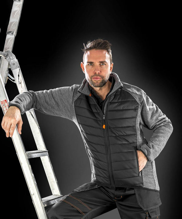 A person with short hair leans against a ladder, wearing a Result R314X gray and black quilted jacket and black pants, with a black background.