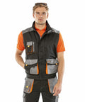 A man wearing an orange shirt, a Result R317X black and gray utility vest with multiple pockets, and matching utility pants stands facing the camera with a neutral expression. He has sunglasses resting on his head.