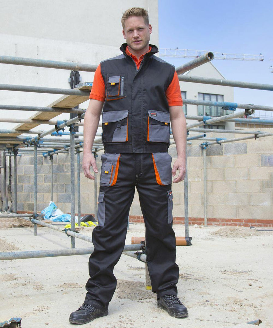 A man stands outdoors wearing a black and orange Result R318X construction uniform with multiple pockets. Scaffolding and building materials are visible in the background.
