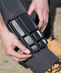 A person adjusts a black Result R322X knee pad over their trousers, wearing brown work boots with orange laces.