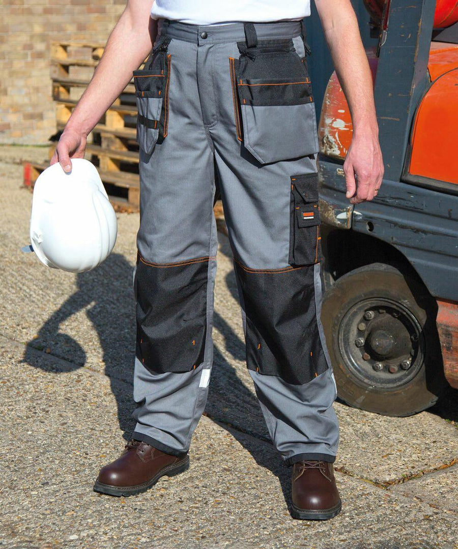 Person wearing gray Result R324X work pants with black patches and brown boots, holding a white hard hat. Standing outdoors near an industrial vehicle.