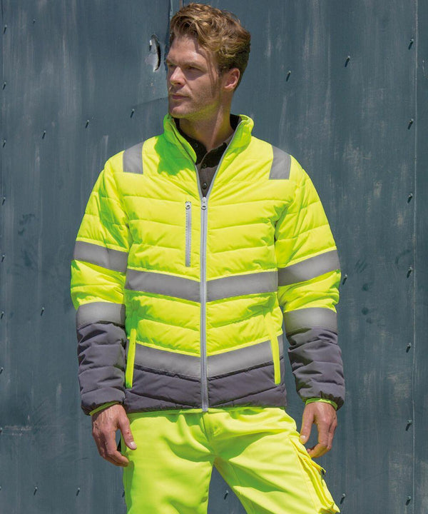 A person wearing a Result R325M high-visibility yellow jacket and pants with reflective stripes stands against a gray industrial background.