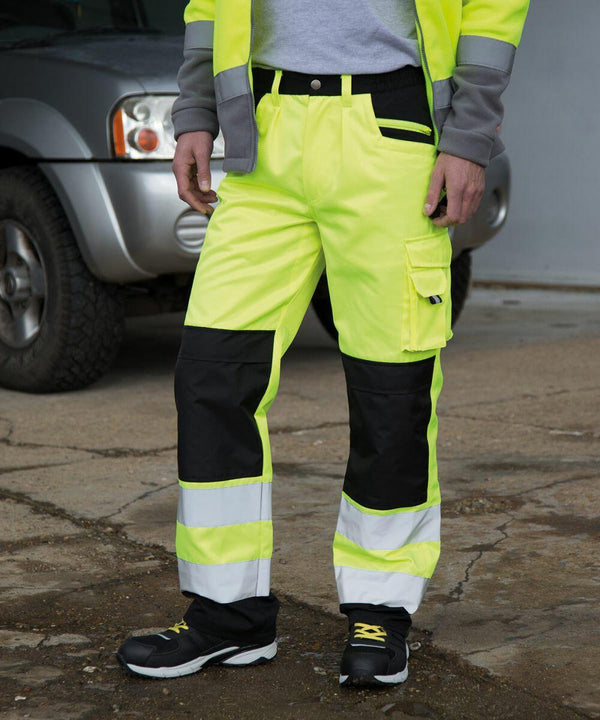 Person wearing Result R327X high-visibility yellow work pants with reflective stripes standing outdoors next to a gray vehicle.