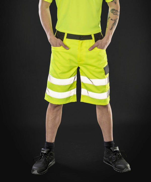 A person wearing bright yellow reflective Result R328X cargo shorts and a matching shirt stands against a dark background. The shorts have black pocket accents and two reflective stripes on each leg. The person also has tattoos.
