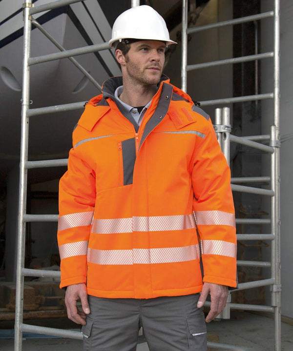 A person wearing a Result R331X orange high-visibility jacket, gray pants, and a white hard hat stands in front of scaffolding.
