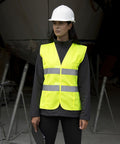 Person wearing a white hard hat and a Result R334F neon yellow safety vest stands in a construction area holding a clipboard.